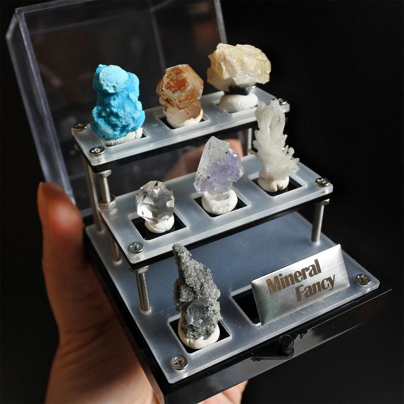 May include: A two-tiered display case with a black base and clear acrylic shelves. The case holds a collection of seven colourful mineral specimens, including a clear quartz crystal, a purple amethyst, a blue turquoise, a white calcite, a brown and orange mineral, and two grey minerals. The case has a silver label that reads "Mineral Fancy".