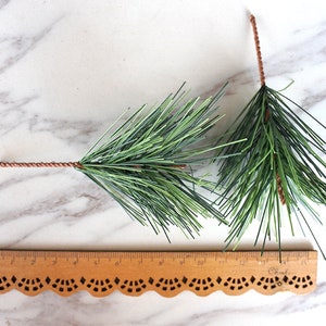 May include: Two sprigs of artificial pine greenery with brown wire stems. The sprigs are arranged on a white marble surface with a wooden ruler in the foreground.