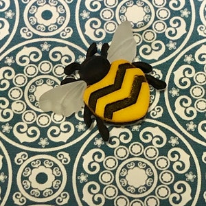 May include: A bee-shaped ornament with a yellow and black striped body, black head, and translucent wings. The bee is set against a teal background with a white circular pattern. This decorative item could be used as a brooch or a decorative element.