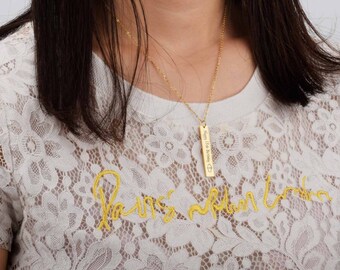 May include: A gold-colored necklace with a rectangular pendant. The pendant has the inscription "Happy 13th Birthday" and a heart symbol. The necklace is worn over a white lace shirt with yellow embroidery.