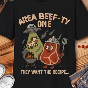 Puede incluir: Camiseta negra con un gráfico humorístico que presenta un personaje de filete de dibujos animados sosteniendo una espátula, una vaca siendo abducida por un OVNI y el texto "AREA BEEF-TY ONE THEY WANT THE RECIPE..."