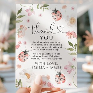 Pode incluir: Um cartão de agradecimento retangular com ilustrações em aquarela de joaninhas, flores cor-de-rosa e folhas verdes. O texto diz: thank YOU for showering our baby with love, and for sharing with us the celebration of our sweet little baby! WITH LOVE, EMILIA + JAMES.
