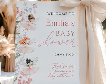Pode incluir: Um cavalete de madeira exibe uma placa de boas-vindas para um chá de bebê. A placa apresenta o texto "Welcome to Emilia's Baby Shower" em rosa, com ilustrações de fadas e flores. Uma grande fita creme adorna a lateral.