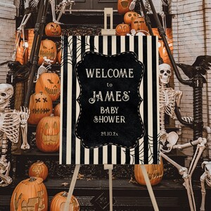 May include: A black and white striped welcome sign with a black chalkboard insert that says "Welcome to James' Baby Shower 24.10.2x". The sign is on an easel in front of a porch decorated with skeletons and pumpkins.