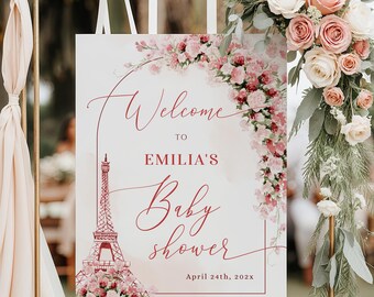 Puede incluir: Un letrero de bienvenida para un baby shower con la ilustración de la Torre Eiffel y el texto "Bienvenida a la Baby Shower de Emilia". El letrero está decorado con flores rosas y se muestra en un soporte dorado.