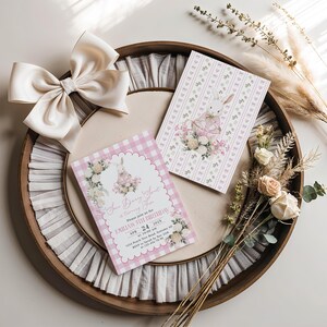 May include: A wooden tray holds two invitation cards with a bunny illustration and floral accents. One card has a pink gingham border, the other a pink and white pattern. A large cream bow and dried flowers complete the arrangement.