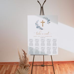 May include: A white seating chart with a gold cross and blue eucalyptus wreath. The chart is titled "Take a Seat" and lists the names of guests for James' First Holy Communion on April 2023.