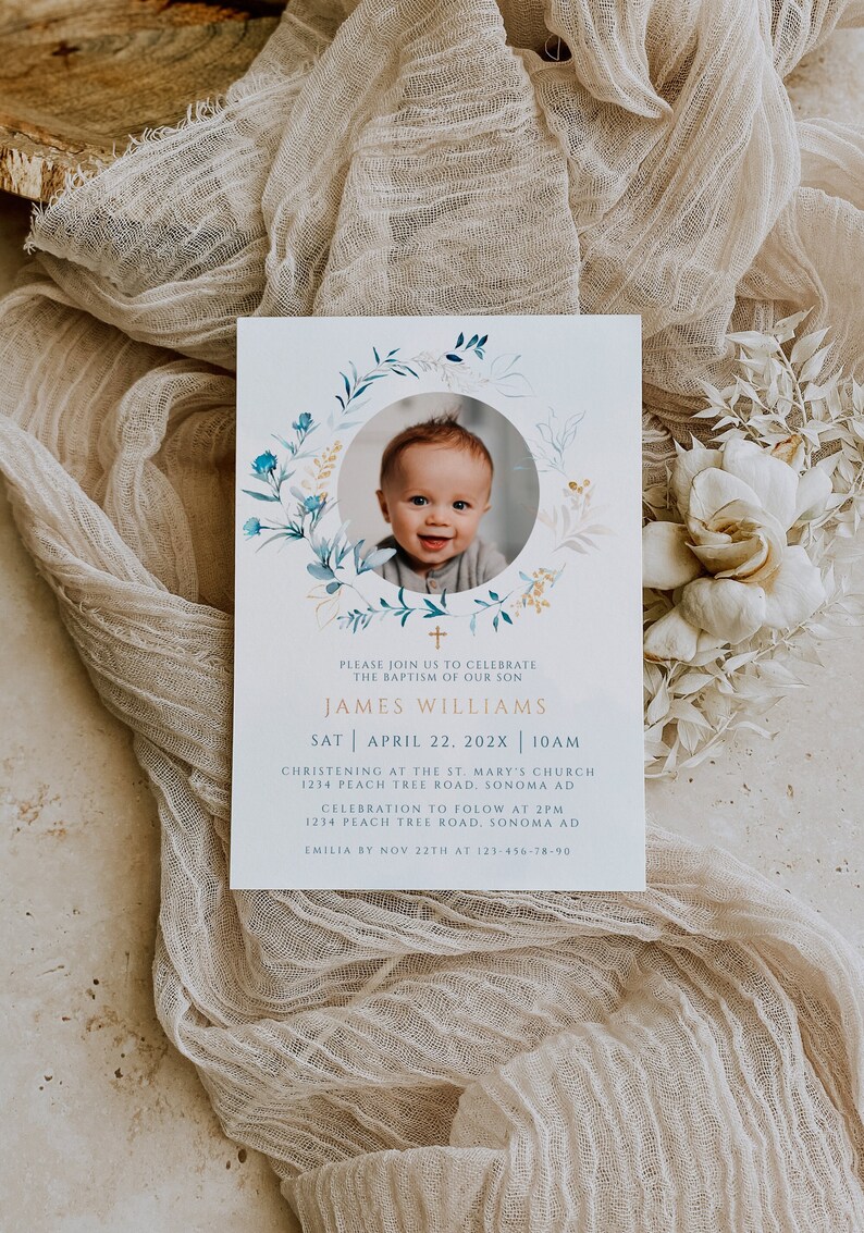 May include: Baptism invitation featuring a smiling baby's photo within a floral wreath. The invitation includes the text "James Williams" and event details: date, time, and location. Soft beige fabric and floral accents complete the design.