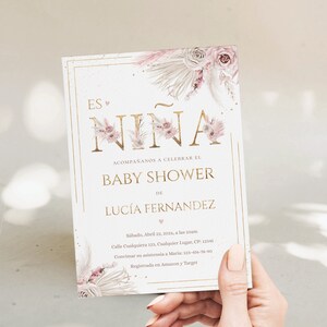 Pu&ograve; includere: Un invito per baby shower con la scritta "Es Ni&ntilde;a" in lettere dorate, decorato con illustrazioni floreali e di piume rosa e beige. L'invito include i dettagli dell'evento, come la data, l'ora e il luogo.