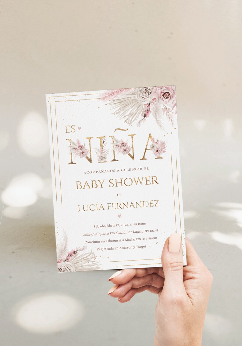 Pu&ograve; includere: Un invito per baby shower con la scritta "Es Ni&ntilde;a" in lettere dorate, decorato con illustrazioni floreali e di piume rosa e beige. L'invito include i dettagli dell'evento, come la data, l'ora e il luogo.