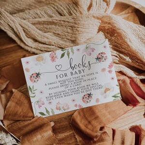 May include: A rectangular baby shower card with the text "books FOR BABY" in a whimsical font. The card is decorated with watercolor flowers and ladybugs. The card is on a wooden surface with tan fabric.