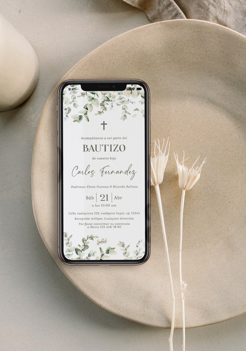 Peut inclure: Un smartphone affiche une invitation de bapt&ecirc;me avec des motifs floraux et le texte "Bautizo" et "Carlos Fernandez". Le t&eacute;l&eacute;phone est pos&eacute; sur une assiette beige, avec des fleurs s&eacute;ch&eacute;es.