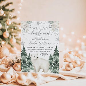 A white invitation card with a winter theme, featuring a polar bear and snowflakes, placed on a table with Christmas ornaments and lights in the background.