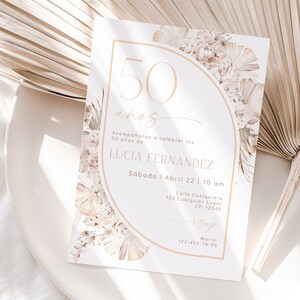 May include: A white rectangular invitation card featuring a large beige arch outline, floral illustrations in neutral tones, and the text 50 a&ntilde;os, Acomp&aacute;&ntilde;anos a celebrar los 50 a&ntilde;os de LUCIA FERNANDEZ, S&aacute;bado | Abril 22 | 10 am, Calle Cualquiera 123.
