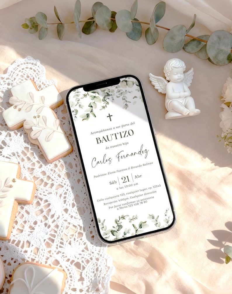 Peut inclure: Un smartphone affiche une invitation de bapt&ecirc;me avec des accents de feuillage vert et le texte "BAUTIZO" et "Carlos Fernandez." Des d&eacute;corations incluent une figurine d'ange et des biscuits en forme de croix. L'invitation est pour le samedi 21 avril.