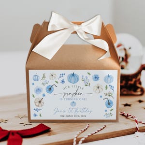 Può includere: Una scatola regalo di cartone marrone con un fiocco di raso bianco. La scatola presenta un'etichetta con zucche blu e bianche, dettagli floreali e il testo "Our Little Pumpkin is Turning One!" e "James' 1st Birthday, September 24th, 202x."