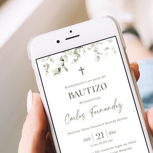 Peut inclure: Un smartphone blanc affiche une invitation de bapt&ecirc;me avec une croix et un motif de feuilles d'eucalyptus. Le texte de l'invitation comprend le nom "Carlos Fernandez" et les d&eacute;tails de l'&eacute;v&eacute;nement. Le t&eacute;l&eacute;phone est tenu dans une main.