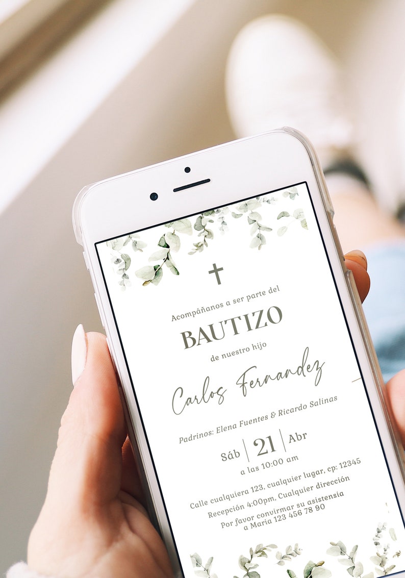 Peut inclure: Un smartphone blanc affiche une invitation de bapt&ecirc;me avec une croix et un motif de feuilles d'eucalyptus. Le texte de l'invitation comprend le nom "Carlos Fernandez" et les d&eacute;tails de l'&eacute;v&eacute;nement. Le t&eacute;l&eacute;phone est tenu dans une main.