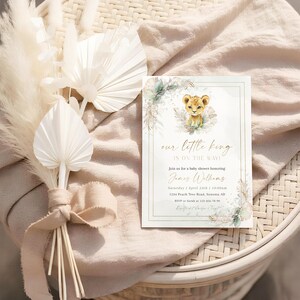 以下が含まれることがあります： 「our little king is on the way!」のテキストが入ったベビーシャワーの招待状。水彩画のライオンの赤ちゃんのイラストが特徴です。招待状は花のアクセントで装飾され、ベージュの布の上に置かれ、装飾用のドライフラワーが添えられています。