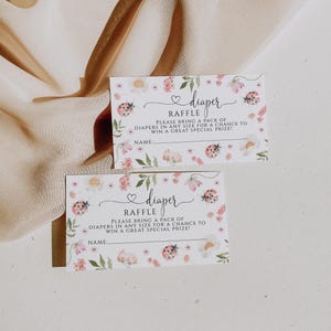 May include: Two baby shower raffle tickets with floral and ladybug designs. The tickets read "diaper raffle" and include space for a name. The tickets are decorated with pink flowers and green leaves.
