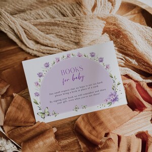 May include: A purple and white floral design card with the text "BOOKS for baby" and a message requesting guests to bring a book in place of a card. The card is on a wooden surface with brown fabric.