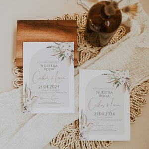 May include: Two rectangular wedding invitation cards featuring a light gray arch graphic, white floral illustrations, and dried palm leaves. The text reads: Te invitamos cordialmente a Nuestra Boda, Carlos & Lucia, 21.04.2024, S&aacute;bado, a las 10:00 am, Calle Cualquiera 123, Cualquier Lugar, CP 12345, Recepci&oacute;n a seguir, Por favor confirmar su asistencia a Maria 123 456 78 90.