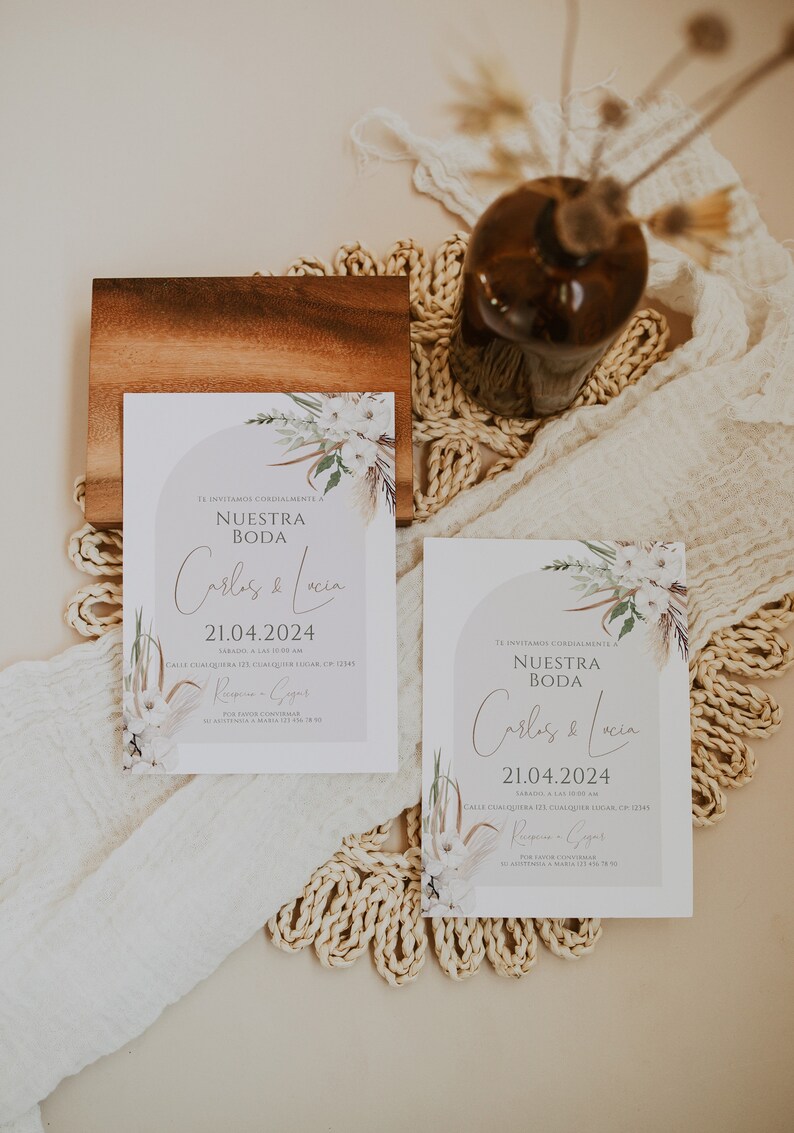 May include: Two rectangular wedding invitation cards featuring a light gray arch graphic, white floral illustrations, and dried palm leaves. The text reads: Te invitamos cordialmente a Nuestra Boda, Carlos & Lucia, 21.04.2024, S&aacute;bado, a las 10:00 am, Calle Cualquiera 123, Cualquier Lugar, CP 12345, Recepci&oacute;n a seguir, Por favor confirmar su asistencia a Maria 123 456 78 90.