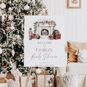 May include: A white sign on a wooden easel with the text "Welcome to Emilia's Baby Shower" in elegant script. The sign features a watercolor illustration of a fireplace, gifts, and a nutcracker. A Christmas tree and gifts are in the background.