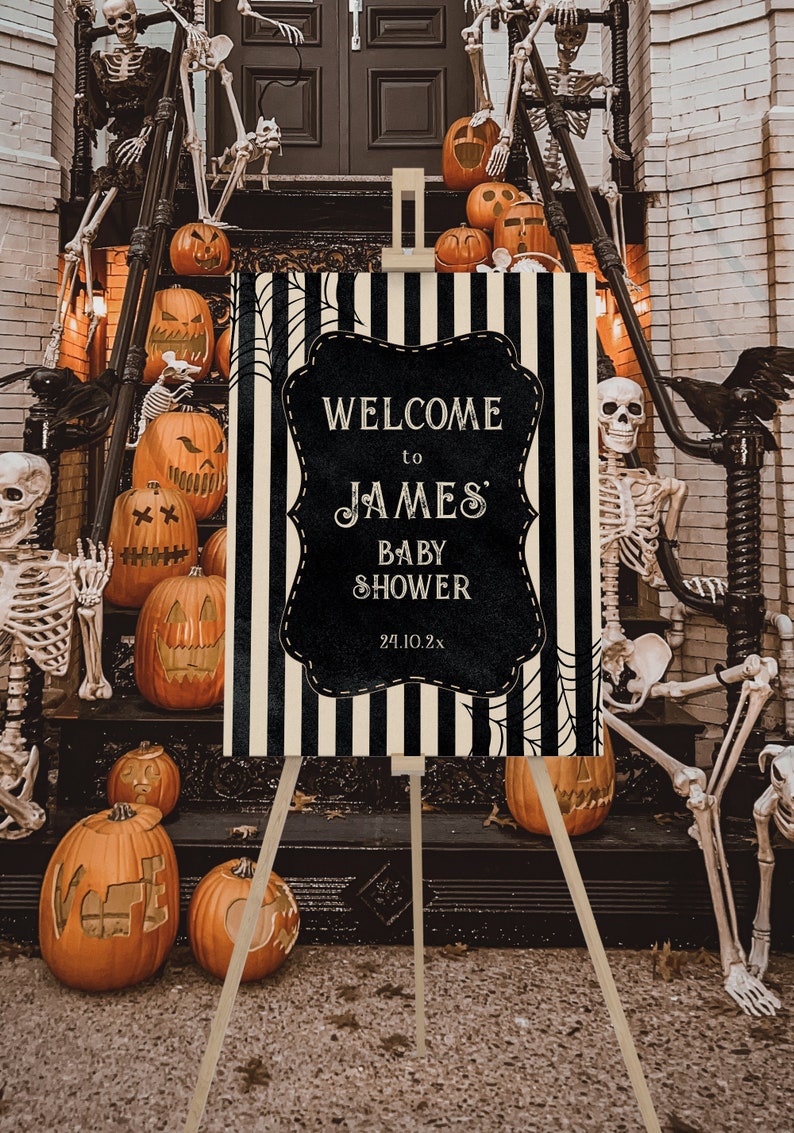 May include: A black and white striped welcome sign with a black chalkboard insert that says "Welcome to James' Baby Shower 24.10.2x". The sign is on an easel in front of a porch decorated with skeletons and pumpkins.