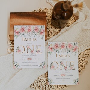 May include: Two rectangular birthday invitation cards featuring a watercolor floral border of pink roses and green leaves. The text reads Emilia is turning one, with event details including the date April 22, time 10:00AM, and location 1234 Peach Tree Road.