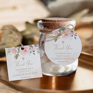 May include: A small glass jar with a cork lid filled with white marshmallows. A round, white tag with pink flowers and green leaves is tied to the jar with twine. The tag has gold text that reads "Thank You for celebrating with us Emilia's Confirmation April 22nd, 202X".