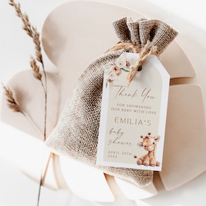May include: A brown burlap bag with a white tag tied to it. The tag features a floral design and the text "Thank You for showering our baby with love Emilia's baby shower April 24th, 202x". A cute giraffe illustration is on the tag.