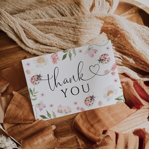 May include: A white card with the words "thank you" in elegant script, adorned with watercolor flowers and ladybugs. The card is surrounded by brown fabric and ribbons, creating a rustic and natural aesthetic.