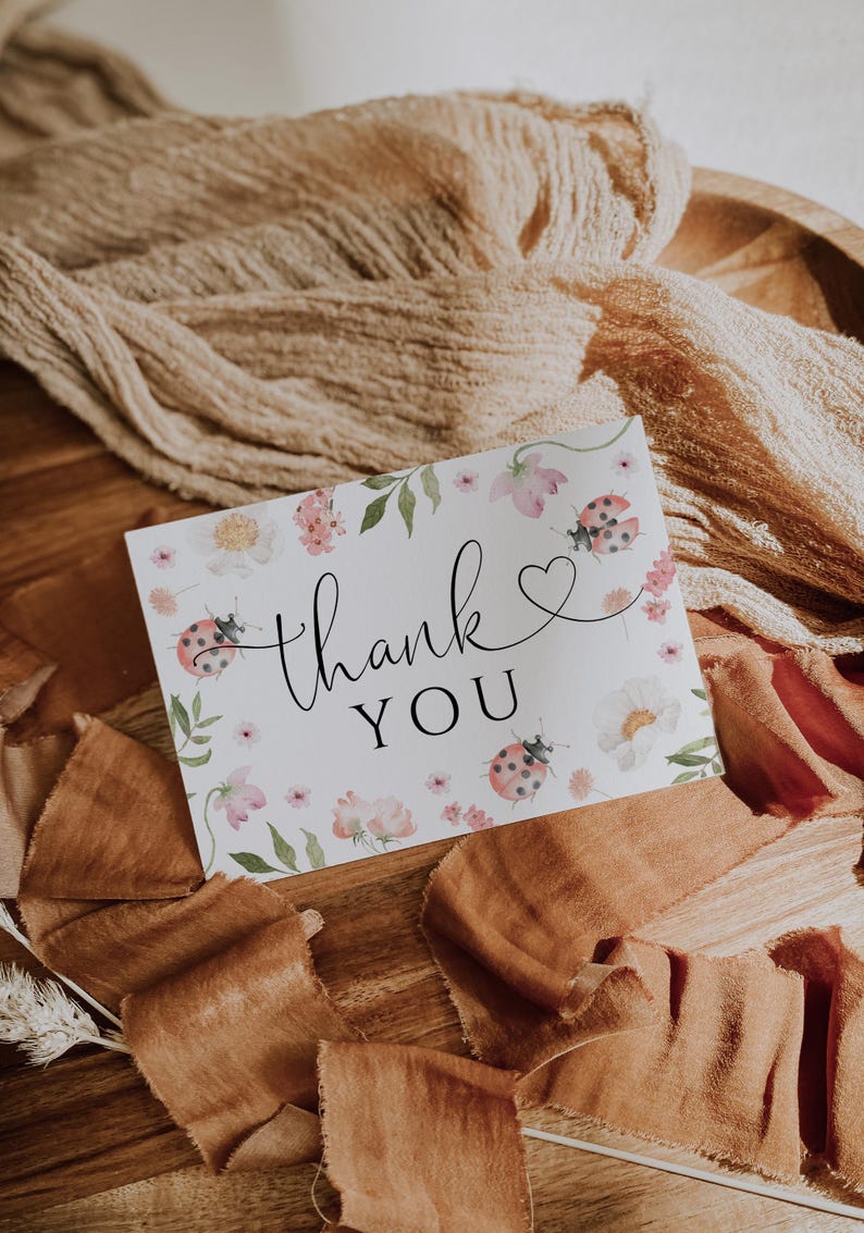 May include: A white card with the words "thank you" in elegant script, adorned with watercolor flowers and ladybugs. The card is surrounded by brown fabric and ribbons, creating a rustic and natural aesthetic.