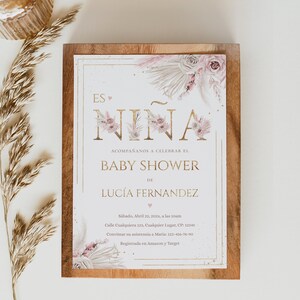 Pu&ograve; includere: Invito per baby shower con la scritta "Es Ni&ntilde;a" in lettere dorate, decorazioni floreali e il nome Luc&iacute;a Fernandez. L'invito &egrave; su uno sfondo di legno con un rametto di erba secca.
