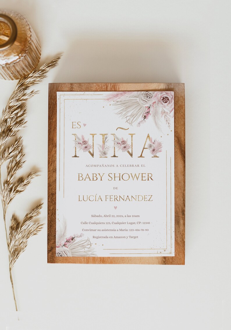 Pu&ograve; includere: Invito per baby shower con la scritta "Es Ni&ntilde;a" in lettere dorate, decorazioni floreali e il nome Luc&iacute;a Fernandez. L'invito &egrave; su uno sfondo di legno con un rametto di erba secca.