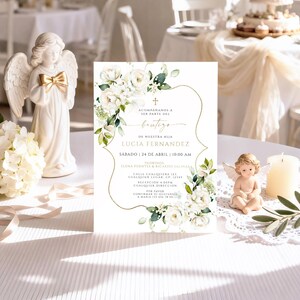 White floral bautizo invitation displayed upright on reception table with angel figurines, candle, and olive branch