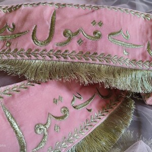 May include: Pink fabric with gold embroidery of Arabic script. The fabric has a gold fringe trim.