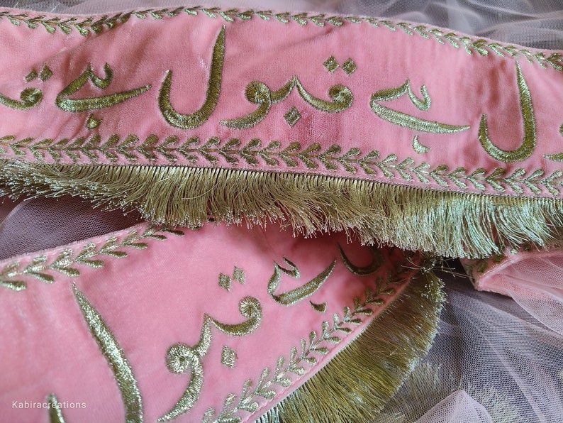 May include: Pink fabric with gold embroidery of Arabic script. The fabric has a gold fringe trim.