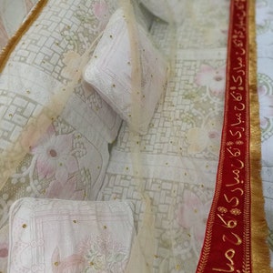 May include: A white bedspread with a sheer overlay and gold trim. The bedspread has a floral pattern and is decorated with gold beads. A red ribbon with gold trim and Arabic text is visible on the right side of the image.