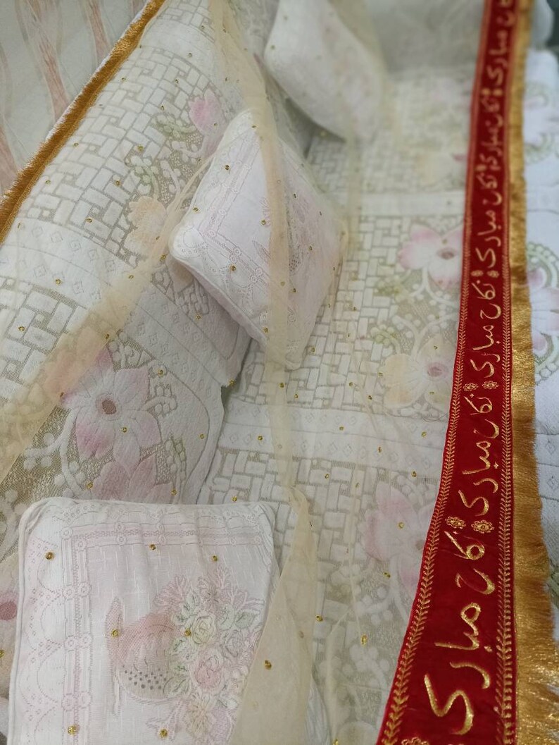 May include: A white bedspread with a sheer overlay and gold trim. The bedspread has a floral pattern and is decorated with gold beads. A red ribbon with gold trim and Arabic text is visible on the right side of the image.
