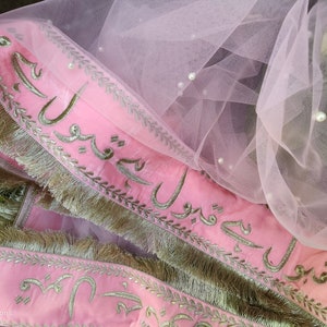 May include: Pink fabric with silver embroidery and fringe trim. The fabric has Arabic script embroidered on it. The fabric is layered over a sheer pink tulle with pearl embellishments.