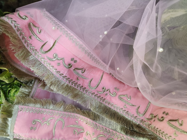 May include: Pink fabric with silver embroidery and fringe trim. The fabric has Arabic script embroidered on it. The fabric is layered over a sheer pink tulle with pearl embellishments.