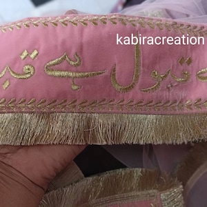 May include: Pink fabric with gold embroidery of Arabic script. The fabric has a gold fringe trim. The text reads "kabiracreation".