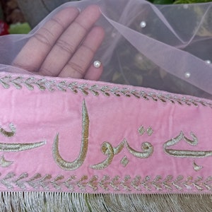 May include: Pink velvet fabric with gold embroidery of Arabic script. The fabric has a silver trim and fringe. The fabric is decorated with small white pearls.