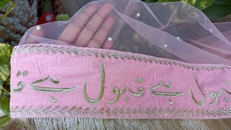 May include: Pink velvet fabric with gold embroidery of Arabic script. The fabric has a silver trim and fringe. The fabric is decorated with small white pearls.