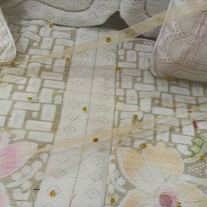 May include: A white fabric with a delicate floral pattern and gold accents. The fabric has a sheer, gauzy texture and is adorned with small, round gold beads.