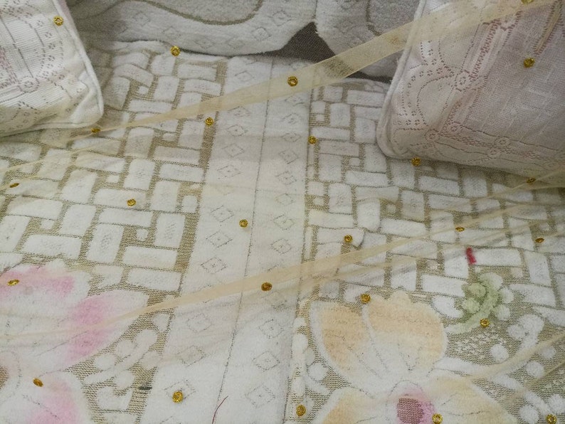 May include: A white fabric with a delicate floral pattern and gold accents. The fabric has a sheer, gauzy texture and is adorned with small, round gold beads.