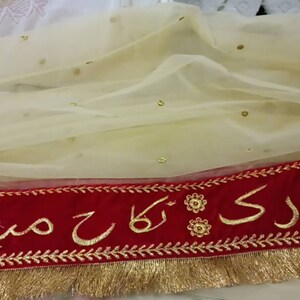 May include: A cream-colored sheer fabric with gold sequins and a red velvet trim. The trim has gold embroidery with the text " Mubarak" in Arabic script.