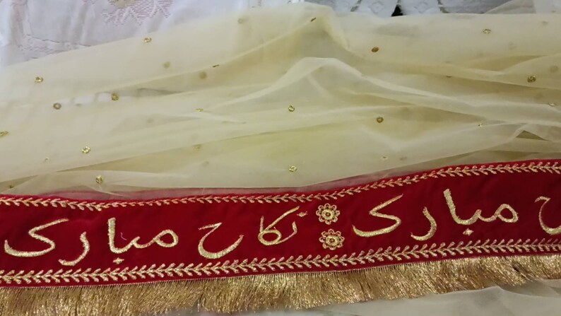 May include: A cream-colored sheer fabric with gold sequins and a red velvet trim. The trim has gold embroidery with the text " Mubarak" in Arabic script.